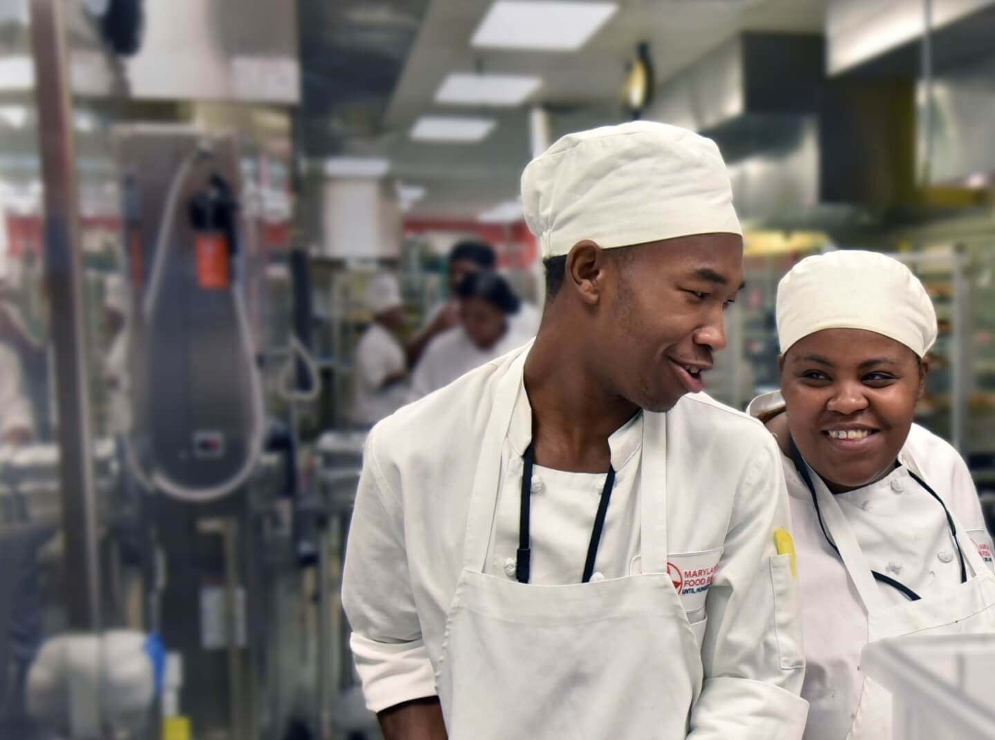Two members of the FoodWorks training program, dressed in chef's gear, smile at each other in the teaching kitchen