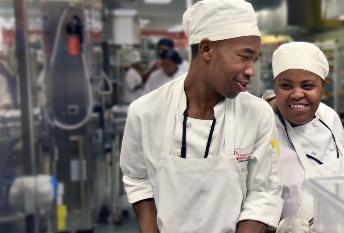 Two members of the FoodWorks training program, dressed in chef's gear, smile at each other in the teaching kitchen