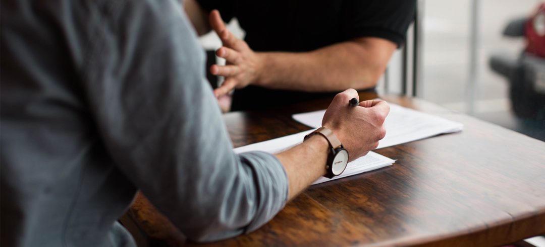 A man prepares to take notes as another man across a small table gestures in conversation.