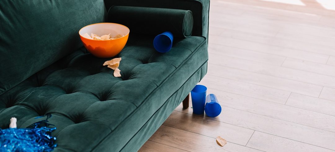 A solitary green couch holding an orange bowl of corn chips, a blue pom-pom, and blue cup.