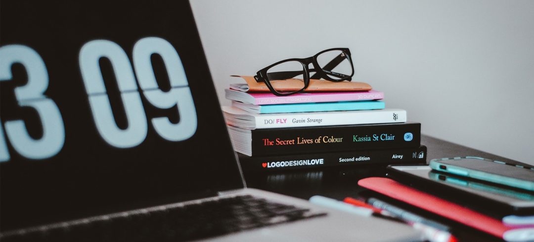 A laptop site open alongside a short pile of books, reading glasses, and a smartphone.
