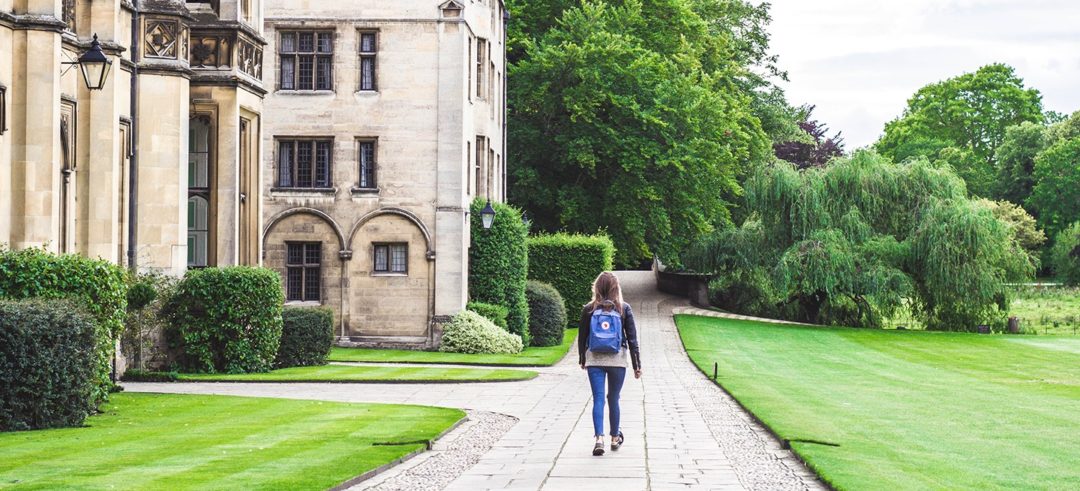 A woman wearing a backpack walks a path alongside a large, gothic university building.