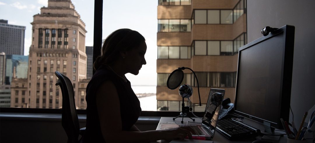 A woman in silhouette sits at her computer desk in front of window displaying a city skyline.