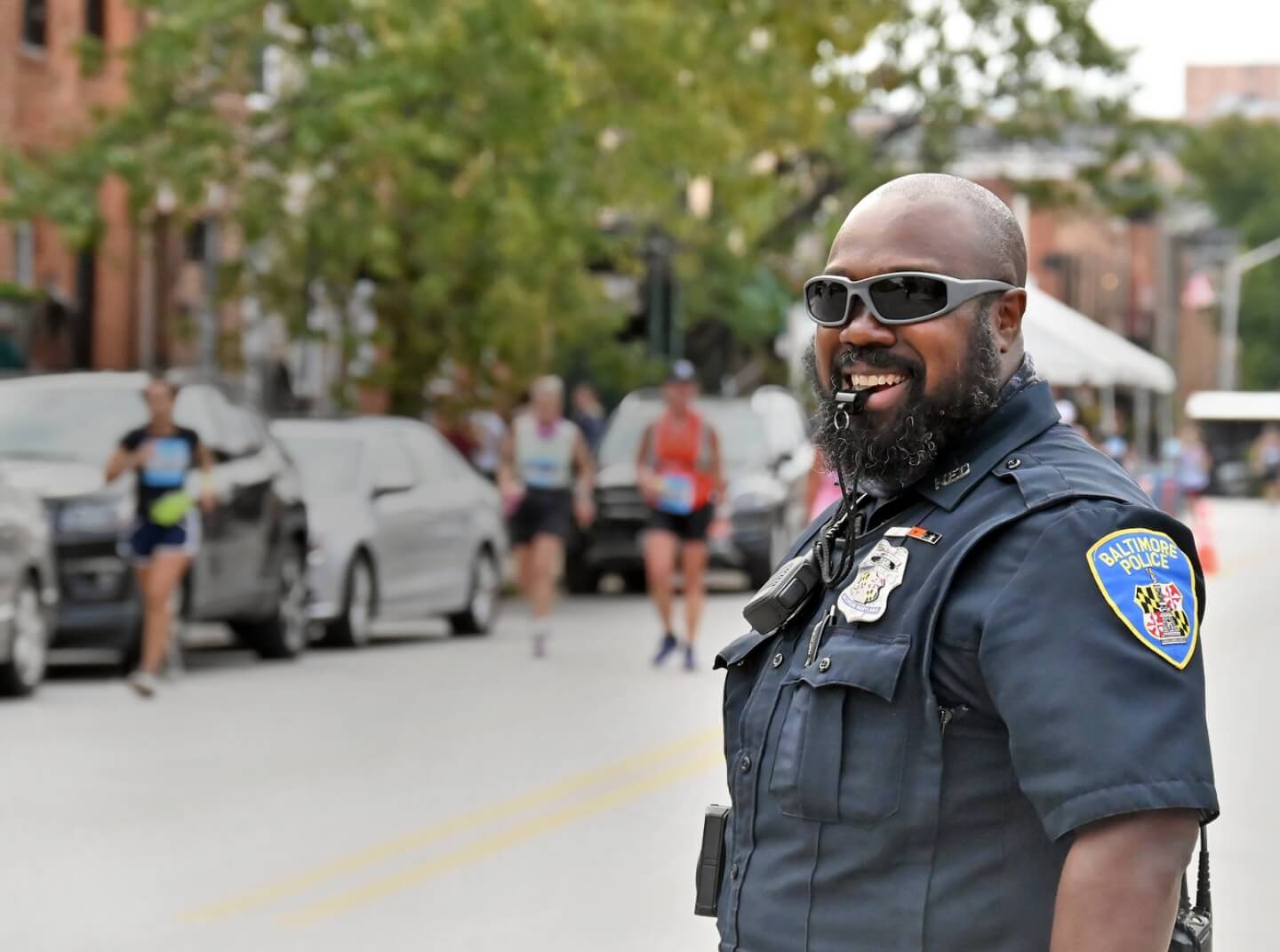 Baltimore Police Officer smiles, wearing sunglasses and holding a traffic control whistle in his mouth