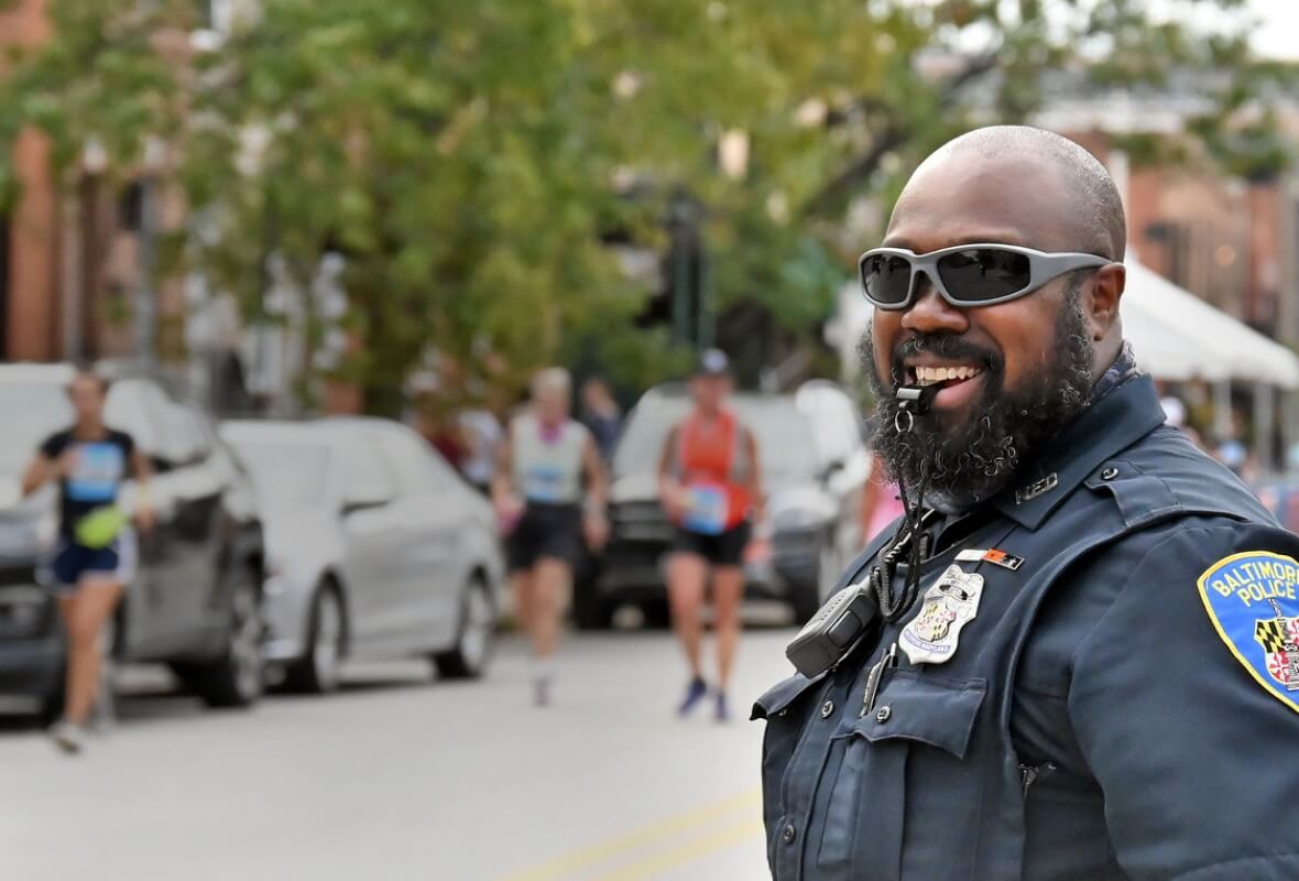 Baltimore Police Officer smiles, wearing sunglasses and holding a traffic control whistle in his mouth