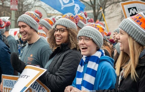 University of Mary students wearing matching knit hats hold signs and cheer at a pep rally