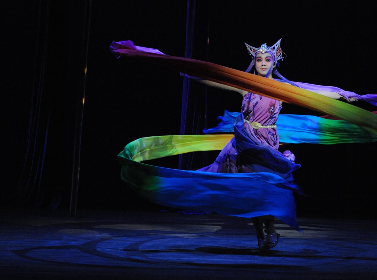 An elaborately costumed dancer twirls during a staged production, causing a long rainbow banner to billow outward into a spiral