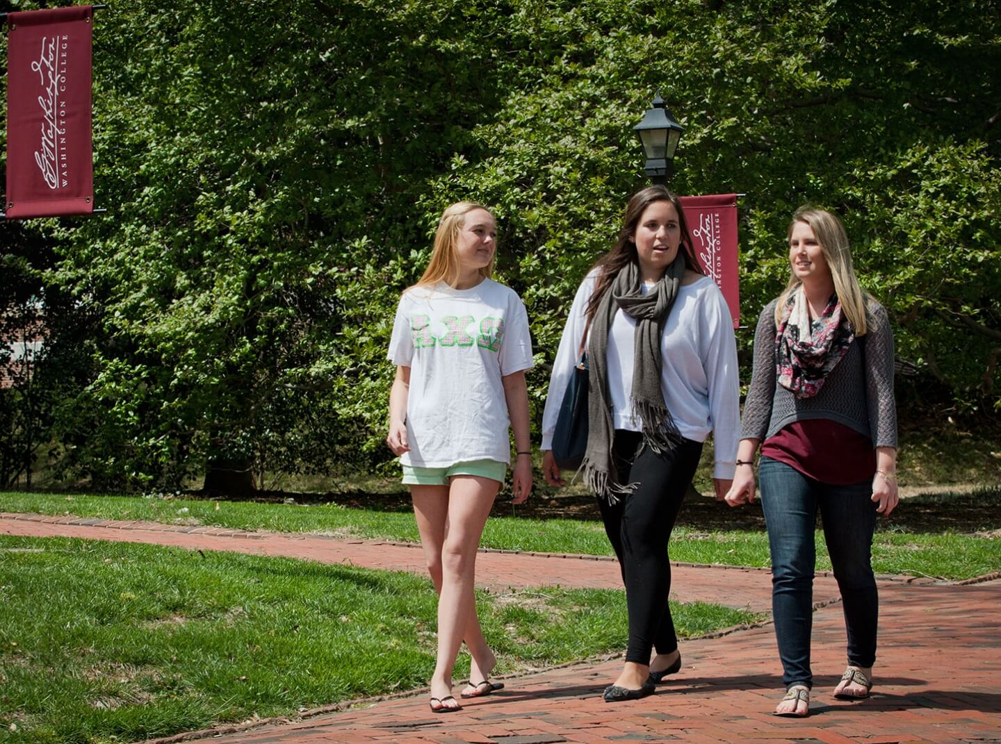 Three Washington College students walk along a brick-paved path on campus during a sunny spring day