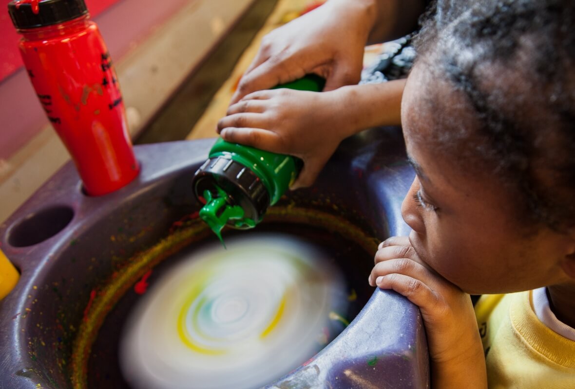 A young girl watches intently as an adult supervisor helps her squeeze paint onto a rapidly spinning card