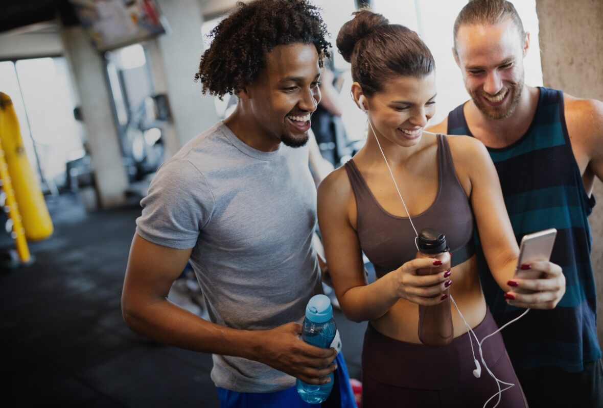 Three gym-going colleagues in workout gear smile at a phone one of them is holding