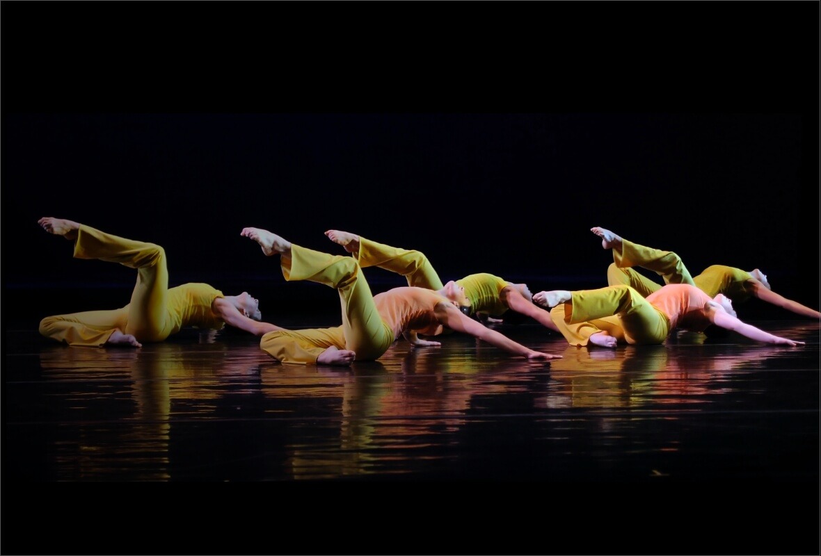 Five students in bright clothing perform a synchronized modern dance, lying on a darkly lit stage with legs extended