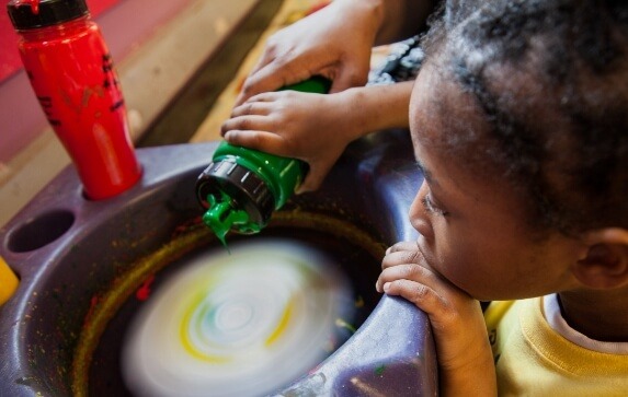 A young girl watches intently as an adult supervisor helps her squeeze paint onto a rapidly spinning card