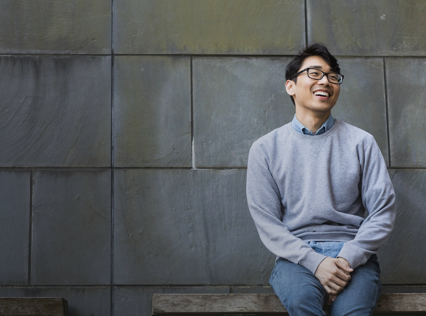 A Drexel Dornsife student wearing glasses smiles while sitting on a bench with a slate wall background