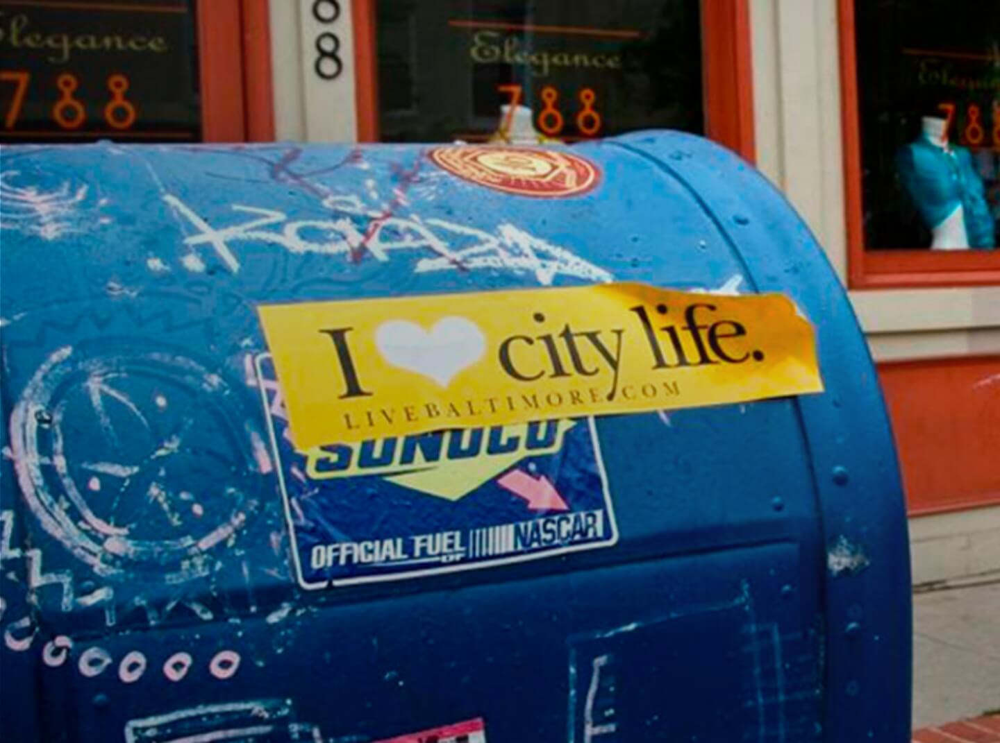 A Baltimore City mailbox with an "I Heart City Life" bumper sticker attached to it