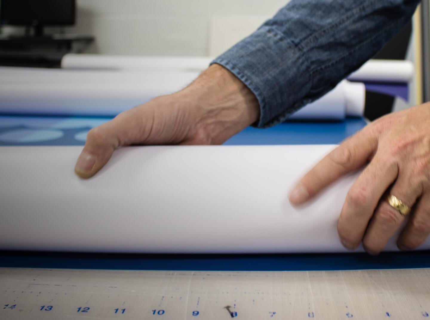 An older man with a wedding ring unrolls a large format print project onto a cutting table