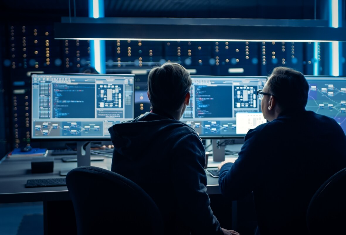 Two IT professionals look toward a data system display across three desktop monitors