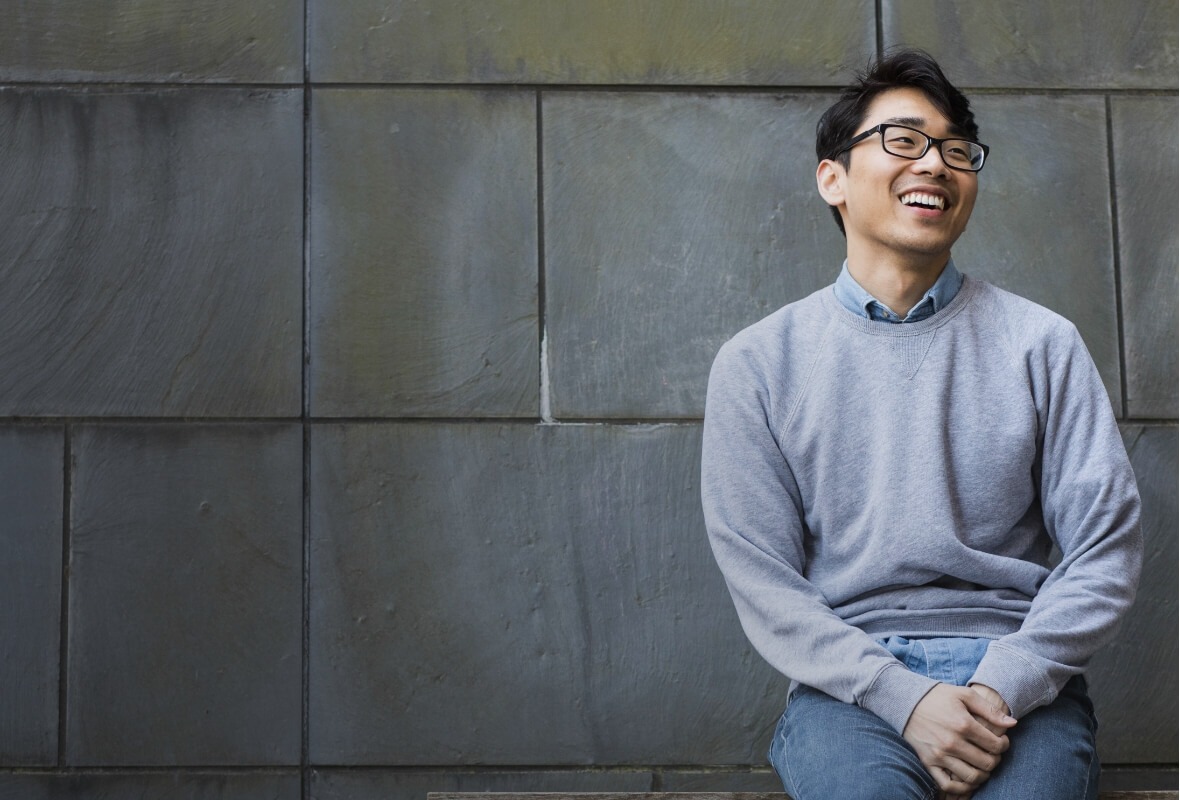 A Drexel Dornsife student wearing glasses smiles while sitting on a bench with a slate wall background