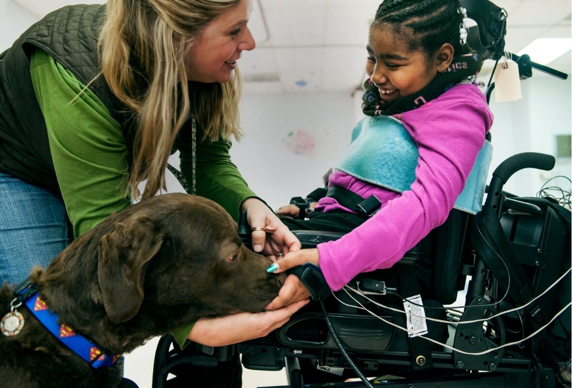 A caretaker introduces a service dog to a young girl in a mechanized wheelchair