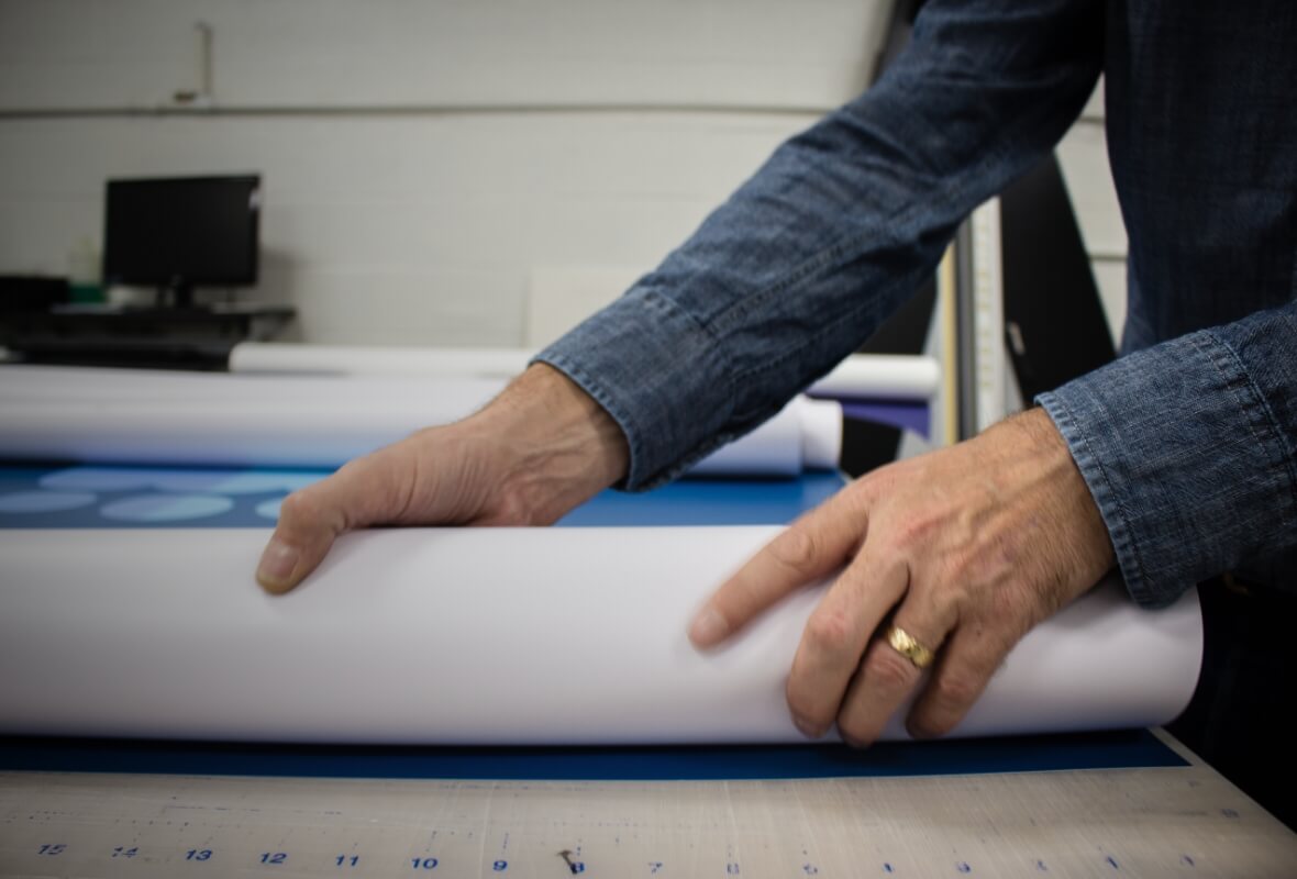 An older man with a wedding ring unrolls a large format print project onto a cutting table