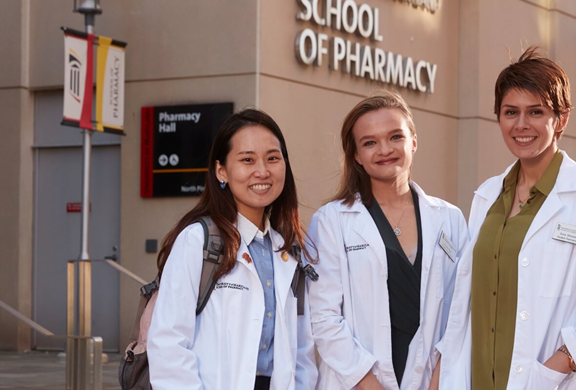 Three University of Maryland School of Pharmacy students wearing lab coats smile while standing in front of a building on campus