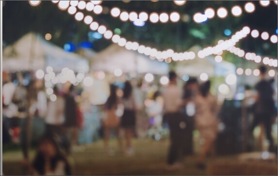 Nighttime outdoor festival decorated with strings hanging lights with a young crowd gathering around vendor tents