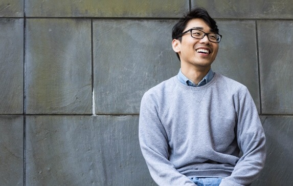 A Drexel Dornsife student wearing glasses smiles while sitting on a bench with a slate wall background