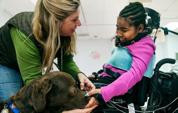 A caretaker introduces a service dog to a young girl in a mechanized wheelchair