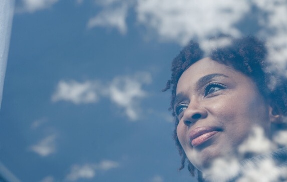 A woman looks out a window which reflects clouds in the sky from the other side