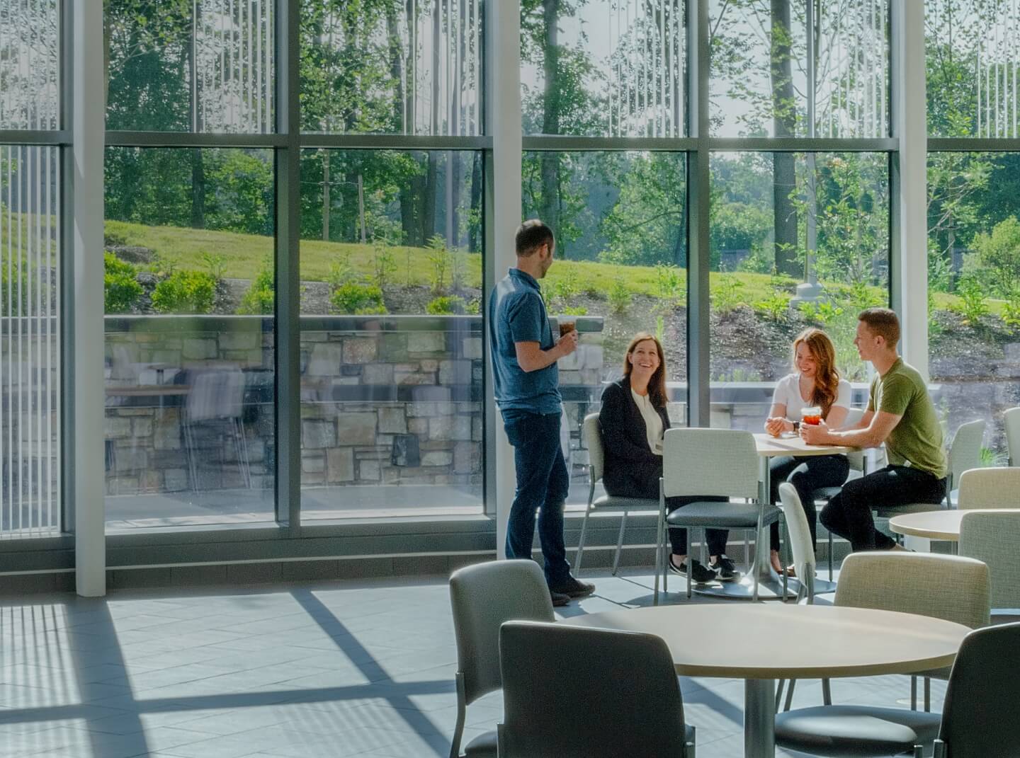 Four young adults chat over coffee at a table in a brightly lit common area with large windows on Sheppard Pratt's new campus