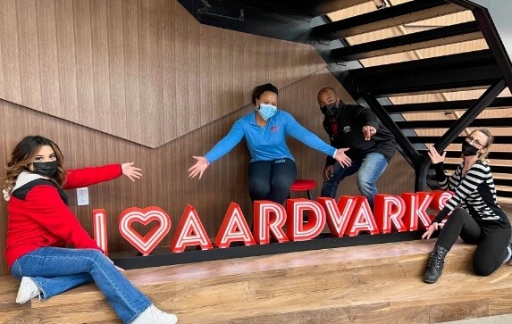 Masked Aims Community College students pose around a neon sign in a campus common area, the sign reads "I heart aardvarks"