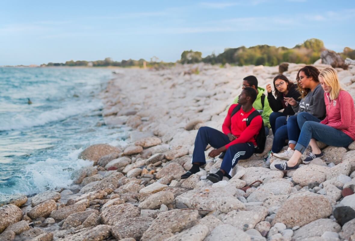 A group of Carthage College students sits on rocks, looking out over Lake Michigan.