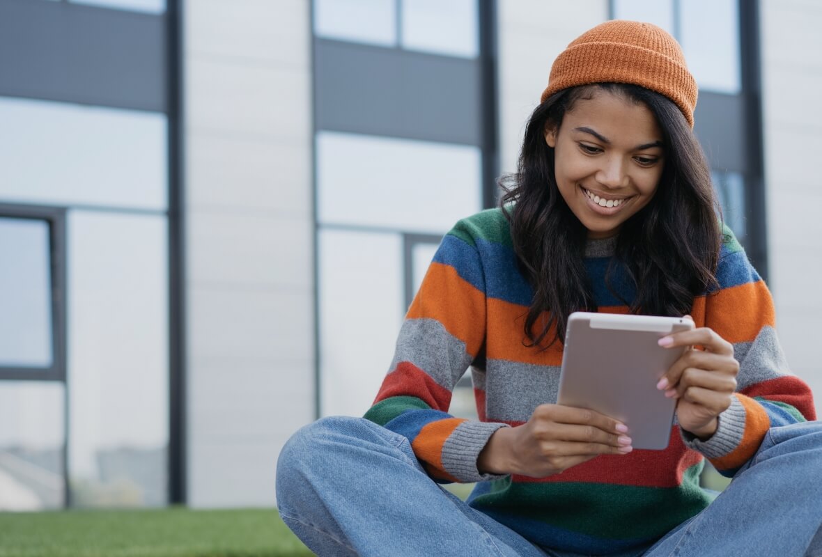 A Howard student in knit hat and sweater sits cross-legged on the ground, smiling and holding a tablet device
