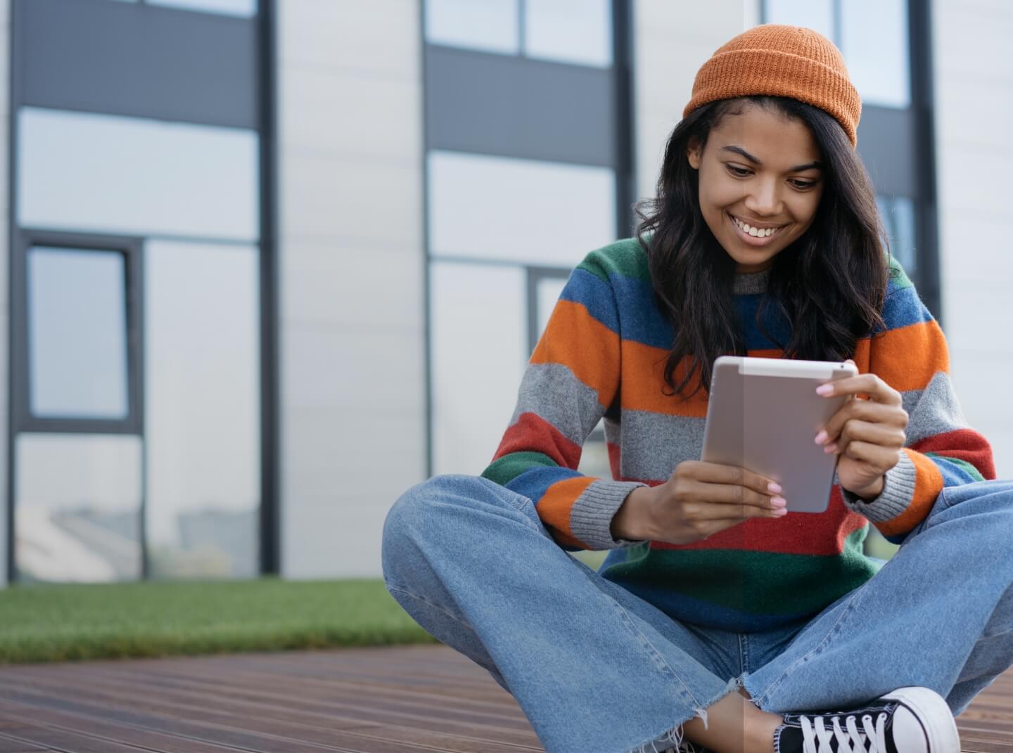 A Howard student in knit hat and sweater sits cross-legged on the ground, smiling and holding a tablet device