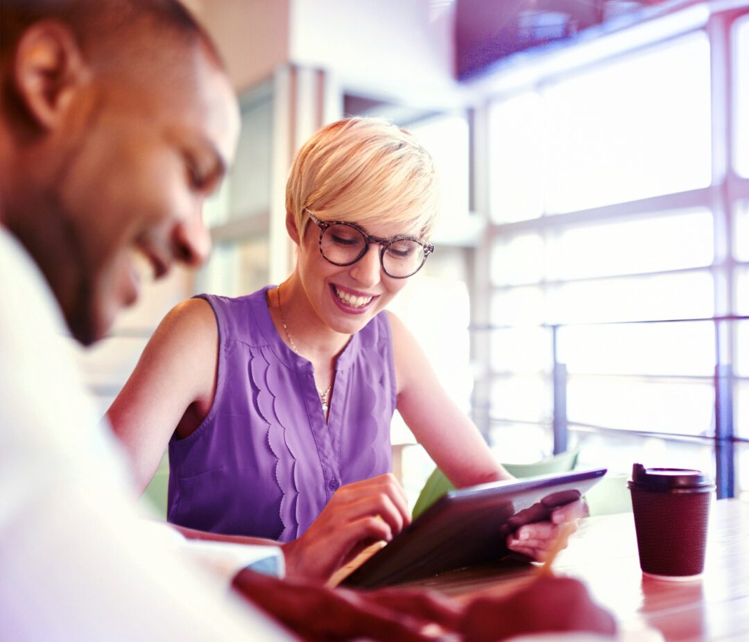 A woman and man sit together at a table in a bright office setting, smiling as they review something on a tablet. The image suggests a collaborative, friendly working relationship and a shared focus on improving ideas or outcomes.