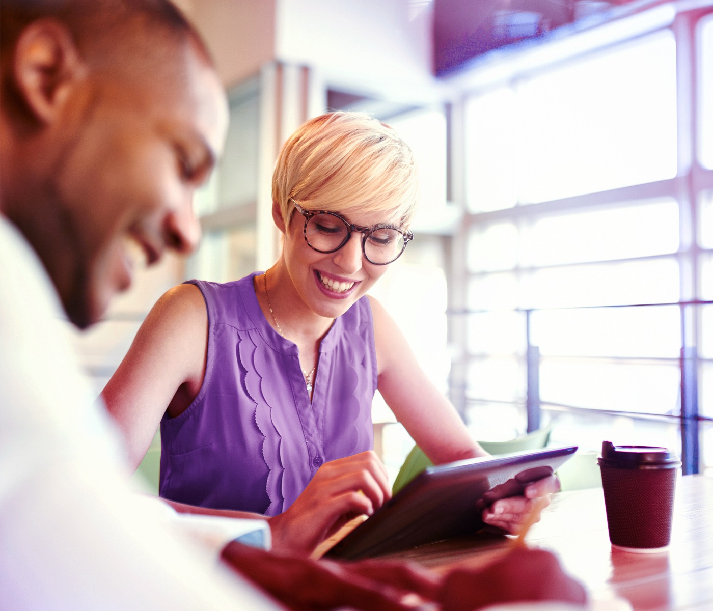 A woman and man sit together at a table in a bright office setting, smiling as they review something on a tablet. The image suggests a collaborative, friendly working relationship and a shared focus on improving ideas or outcomes.